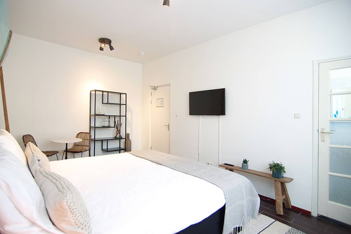 A bedroom with a large bed, a TV mounted on the wall, a small table, and a bench with plants.
