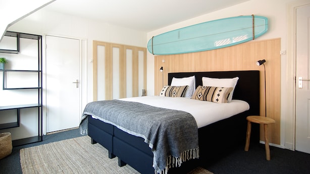 A bedroom with a bed, a surfboard on the wall, a shelf, and a small stool. - Hotel Zand (Zandvoort)