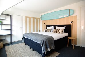 A bedroom with a bed, a surfboard on the wall, a shelf, and a small stool. - Hotel Zand (Zandvoort)