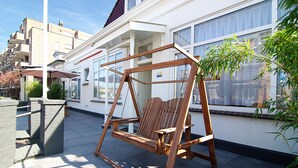 A wooden swing chair in a courtyard with a white building in the background. - Hotel Zand (Zandvoort)