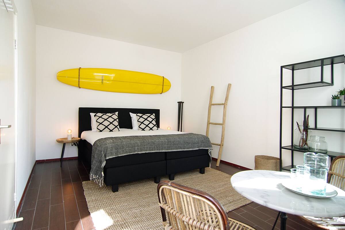 A bedroom with a bed, a bedside table with a candle, a yellow surfboard, a wooden ladder, a wicker chair, and a small table with glasses.