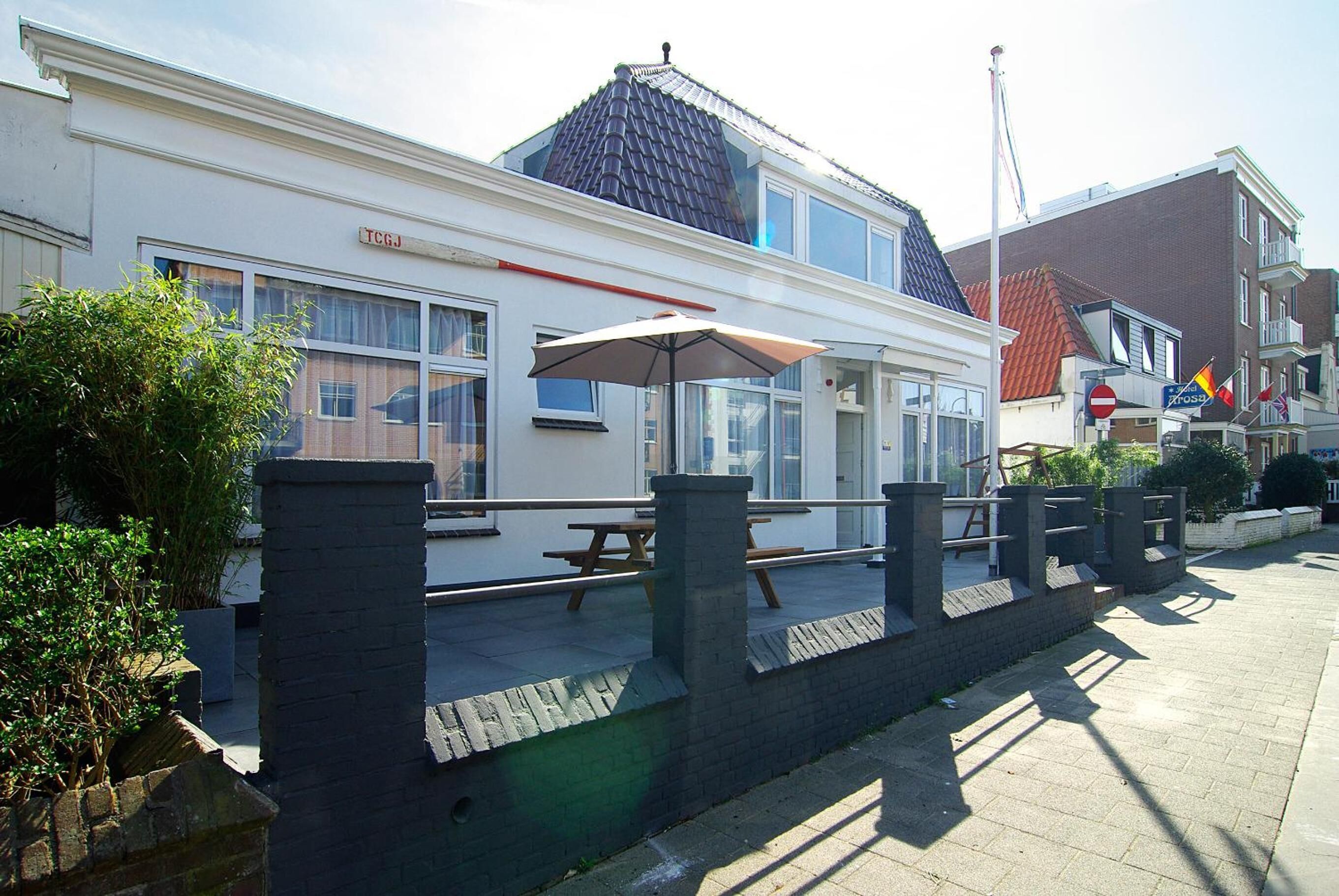 Een wit gebouw met een bord waarop 'TCGJ' staat, en een overdekt terras met een parasol.