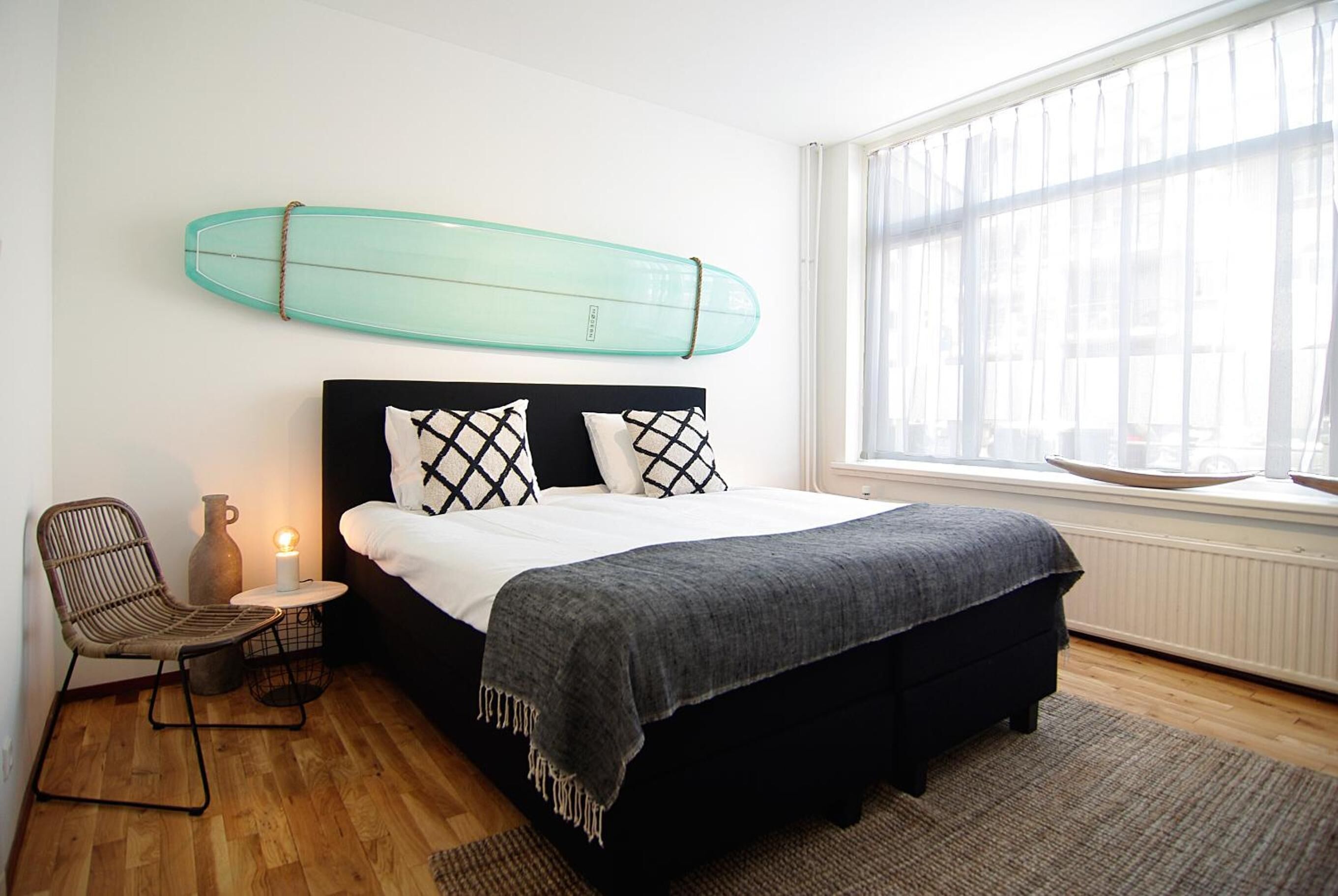 A bedroom with a bed, a chair, a bedside table with a lamp, a surfboard on the wall, and a window with blinds.