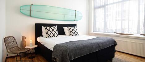 A bedroom with a bed, a chair, a bedside table with a lamp, a surfboard on the wall, and a window with blinds.