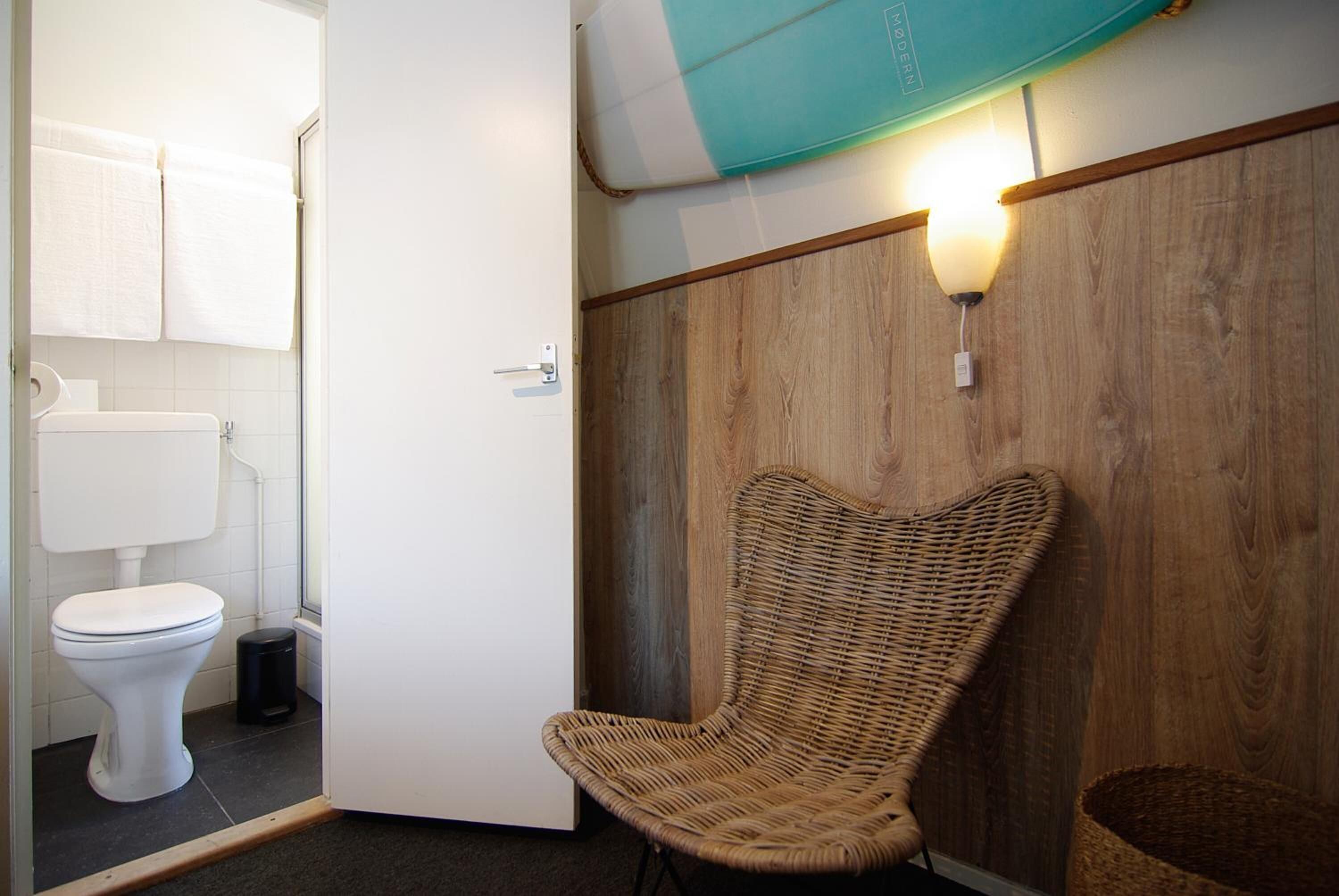 Un baño con inodoro blanco, una silla de mimbre y una pared de madera con una lámpara.