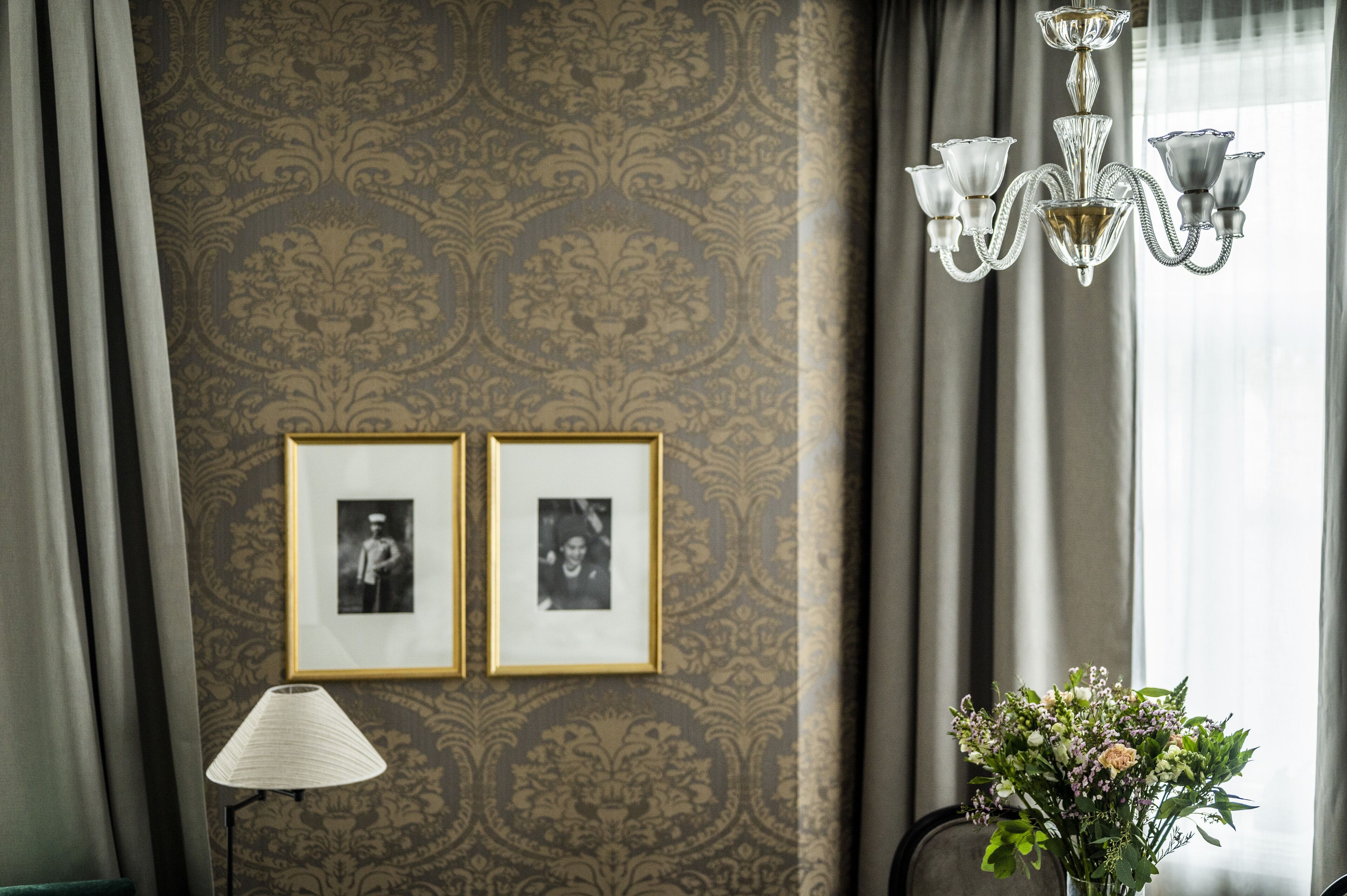 Quarto com papel de parede estampado, duas fotografias emolduradas, um candelabro, uma luminária e um vaso com flores.