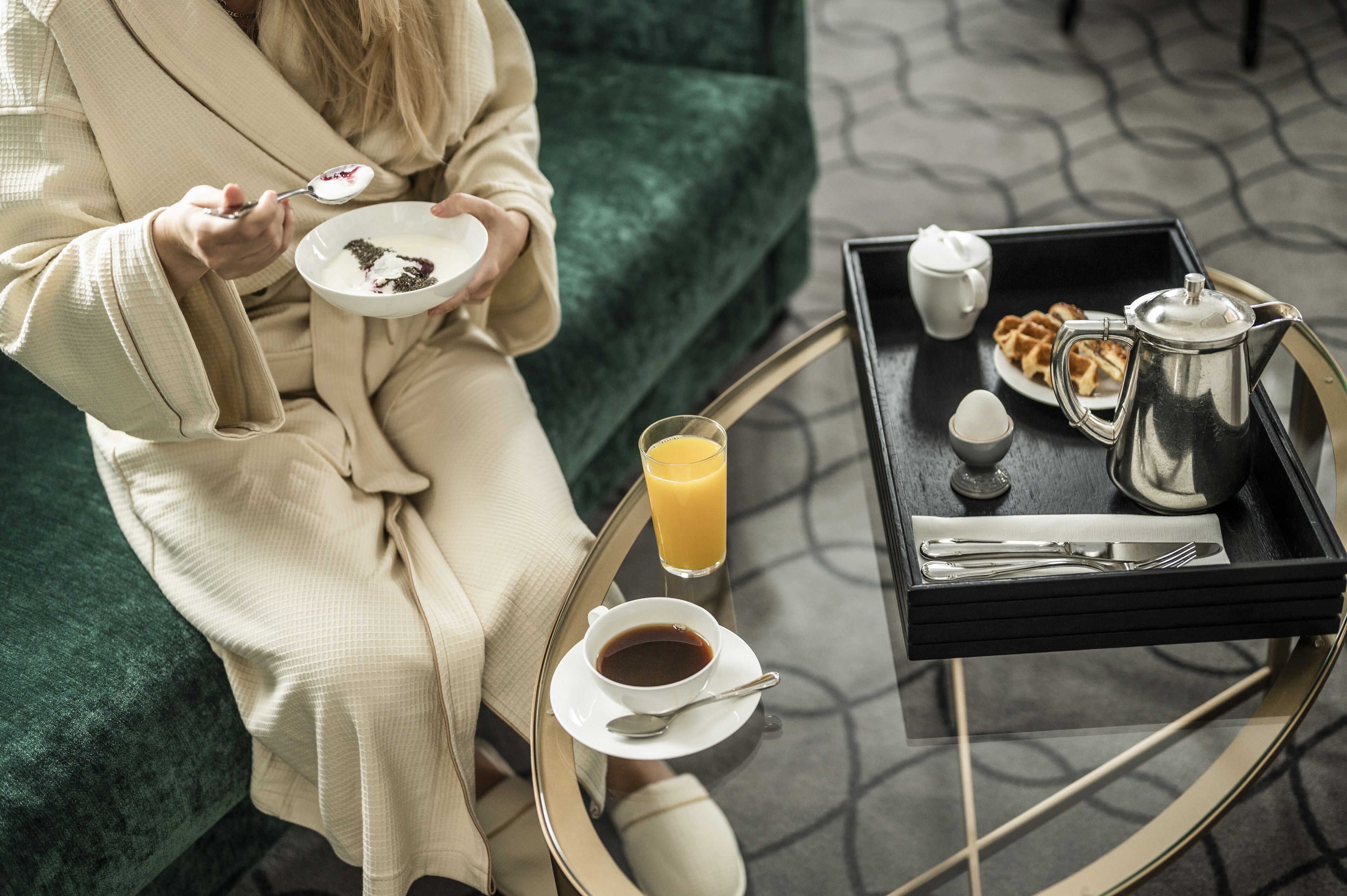A person in a robe holding a plate with a dessert, sitting on a green couch with a tray of breakfast items including waffles, coffee, and juice.