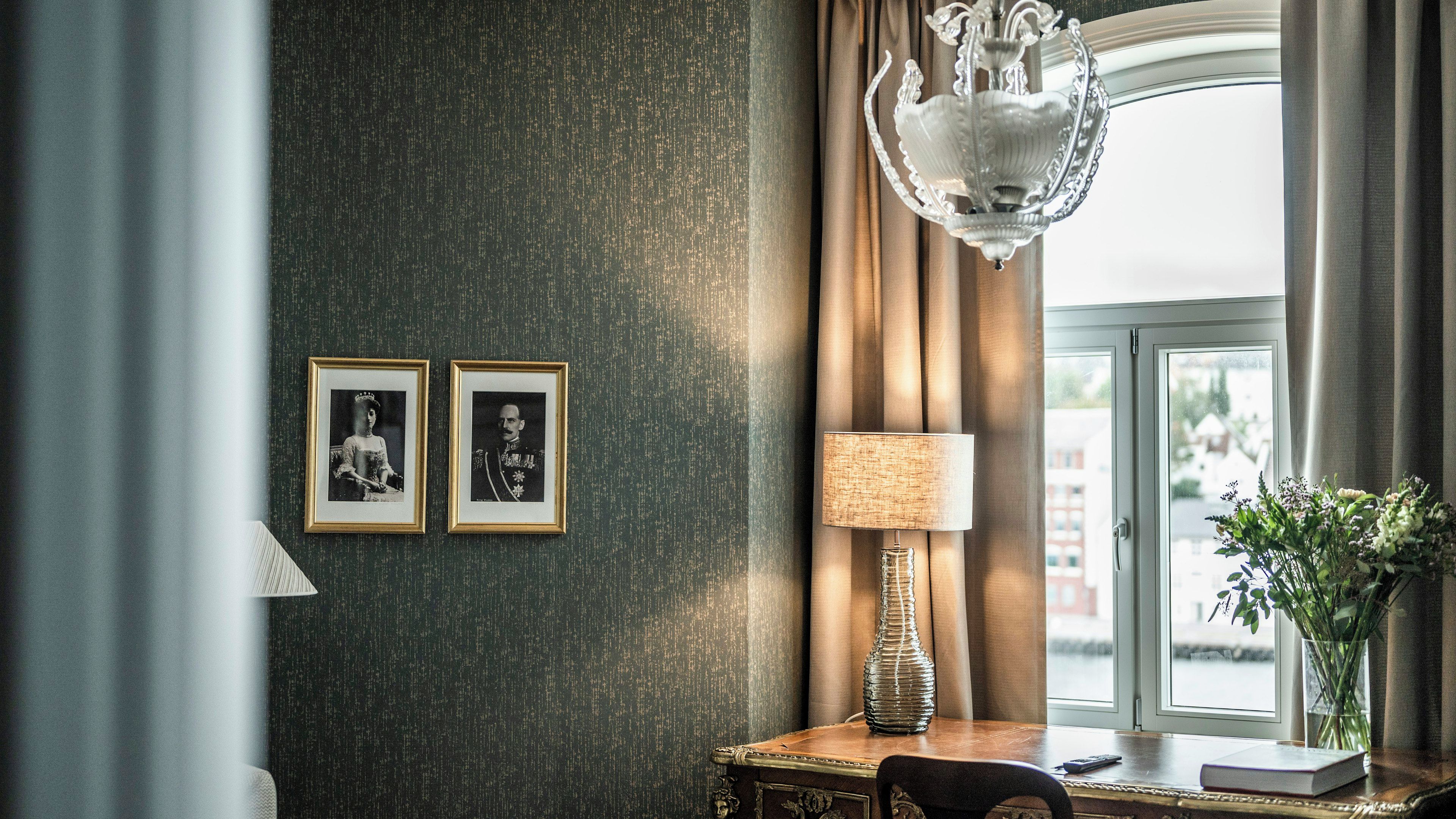 A room with a textured wall, a chandelier, framed photographs, a lamp, a vase with flowers, and a view through a window.