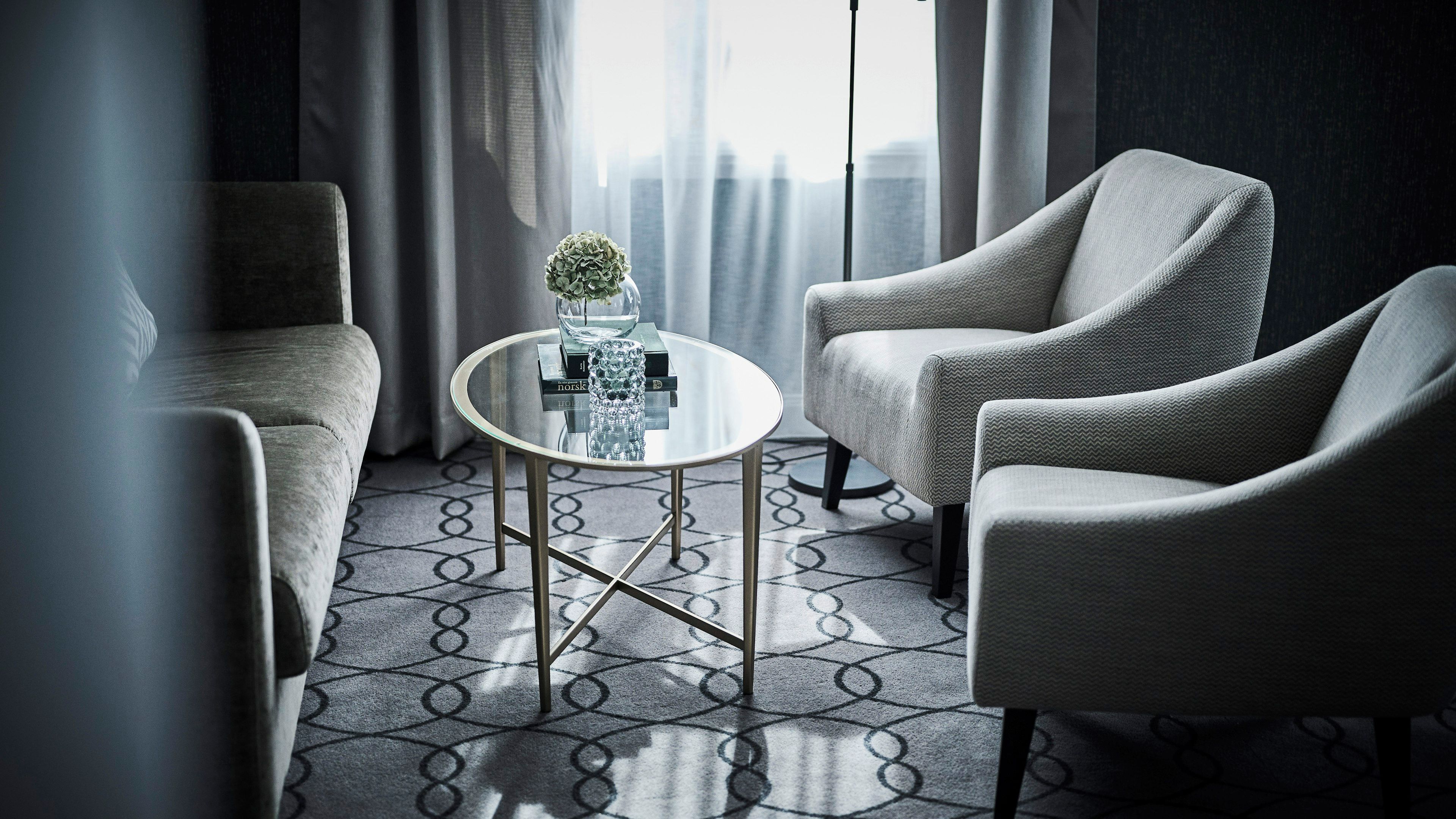 Une chambre d’hôtel moderne avec une table ronde en verre, deux fauteuils, une moquette à motifs et une vue sur les toits de la ville, visible à travers des rideaux transparents.