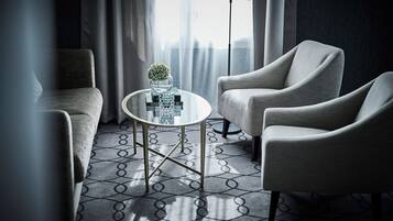 Une chambre d’hôtel moderne avec une table ronde en verre, deux fauteuils, une moquette à motifs et une vue sur les toits de la ville, visible à travers des rideaux transparents.