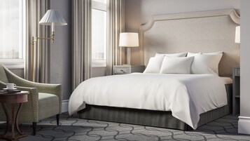Una habitación de hotel moderna con una cama grande, una mesita de noche con lámpara, una silla y vistas a la ciudad desde la ventana.