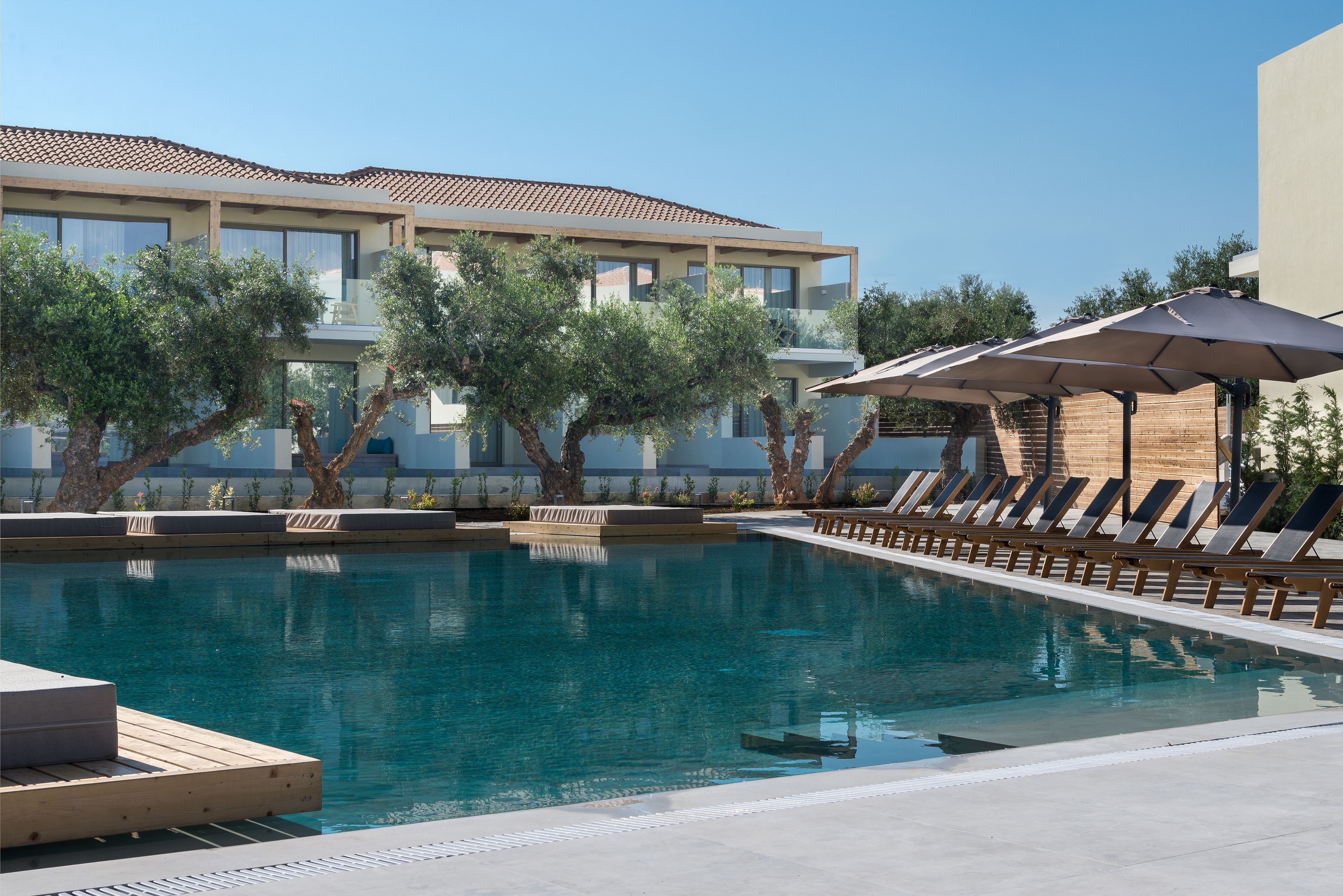 A swimming pool with lounge chairs and umbrellas, surrounded by trees and a building with a tiled roof.