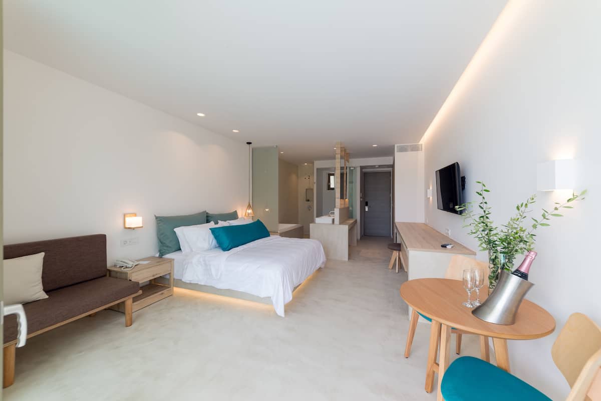 Una habitación de hotel moderna con una cama, mesita de noche, lámpara, televisor y una zona de comedor con mesa y sillas.