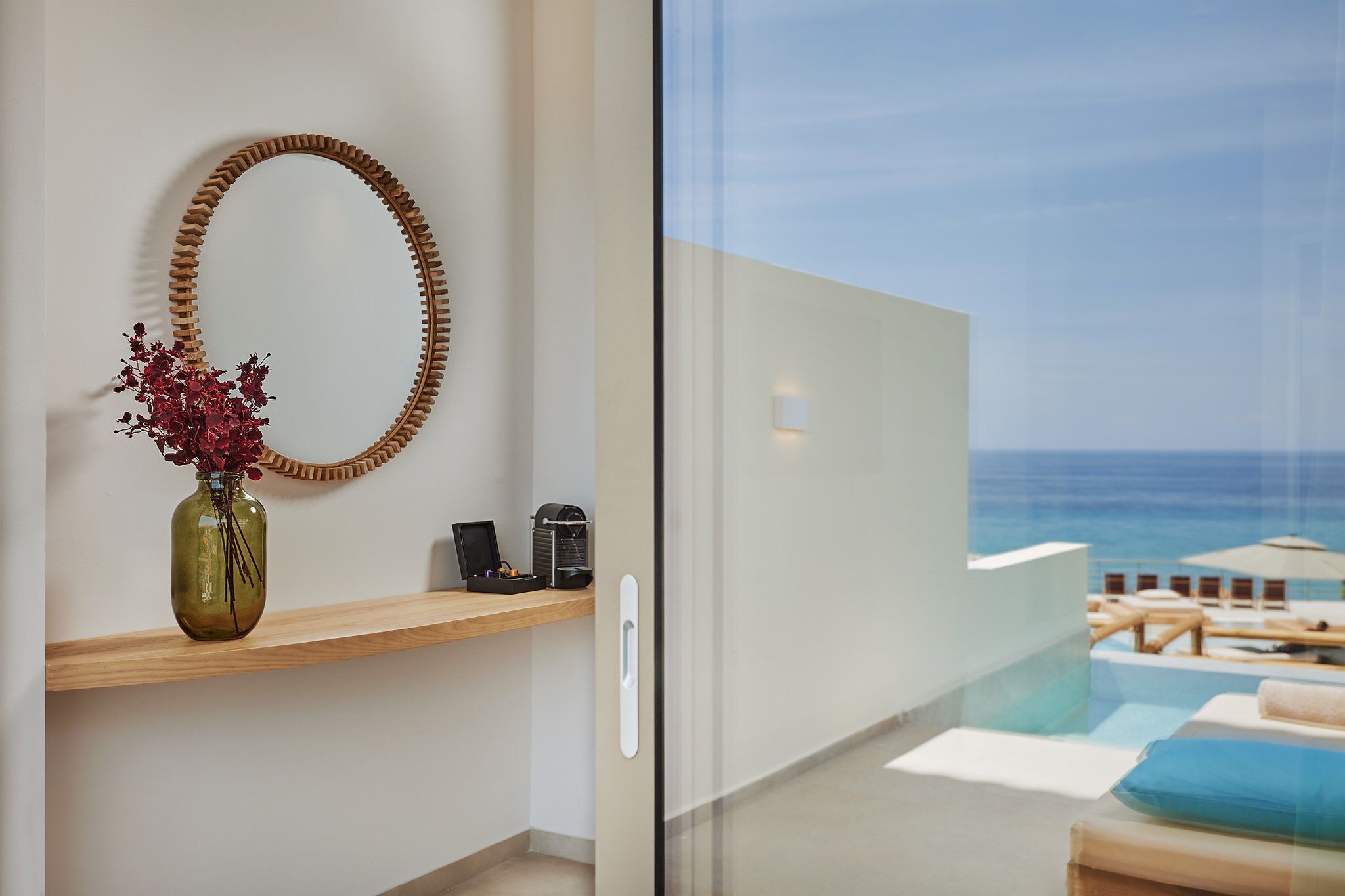 Una habitación de hotel moderna con una estantería de madera que sostiene un florero y un teléfono, una puerta de cristal que da a un balcón con piscina y sillas de descanso.