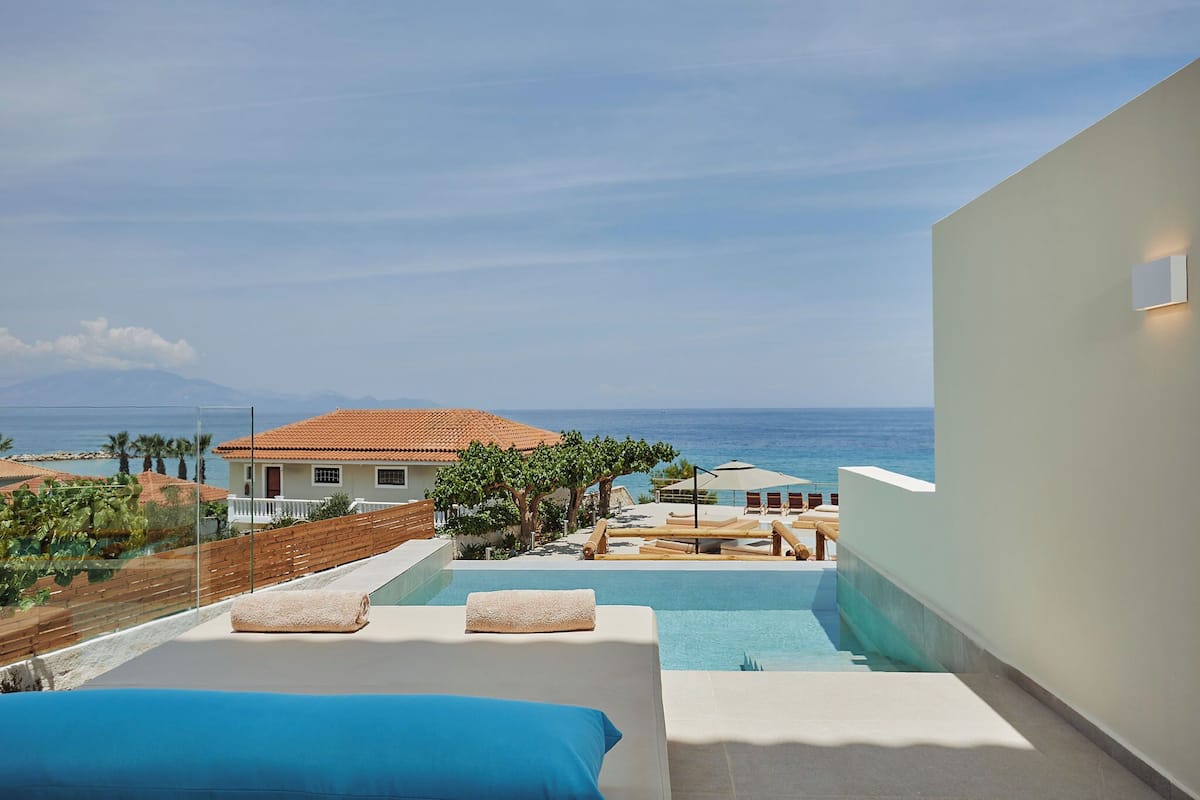 Una piscina infinita en la azotea con vistas al mar y a las montañas, una pared blanca con una luminaria y una terraza de madera con sillones de descanso.