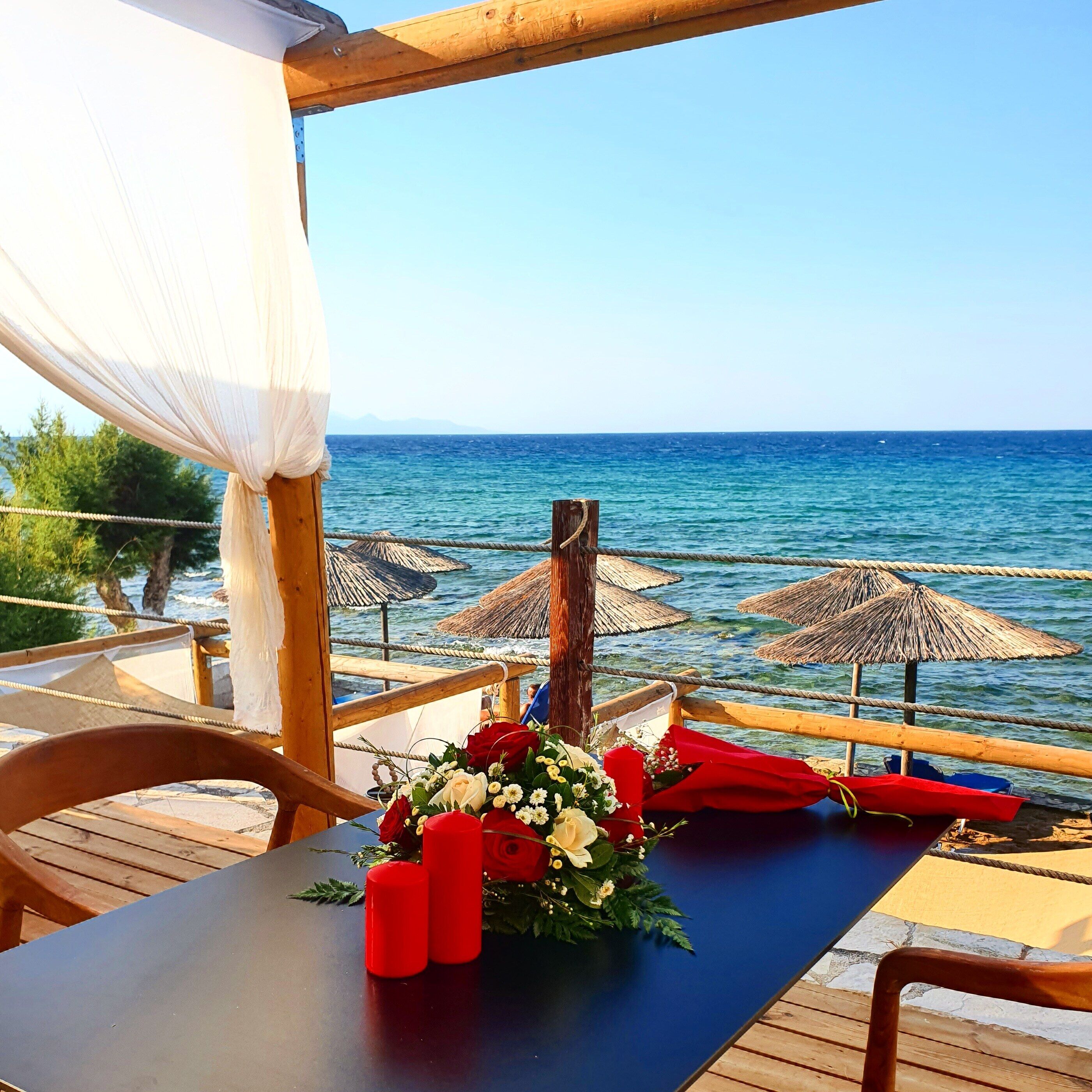 A wooden deck with a table set for dining, overlooking a beach with thatched umbrellas and the sea.