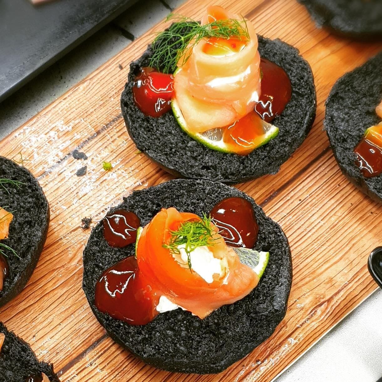 Une planche en bois sur laquelle sont disposés plusieurs morceaux de sushi, posés sur des galettes de riz noir, le tout agrémenté d'herbes vertes et d'une tranche de tomate.
