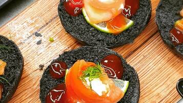Une planche en bois sur laquelle sont disposés plusieurs morceaux de sushi, posés sur des galettes de riz noir, le tout agrémenté d'herbes vertes et d'une tranche de tomate.