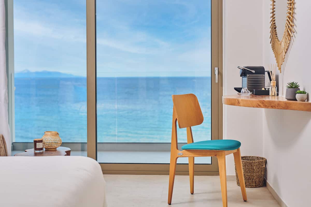 Una habitación con un ventanal grande con vista al mar, una silla de madera con cojín verde azulado, una mesita con una vela y una cesta de mimbre, y un estante con una cafetera y una planta en maceta.
