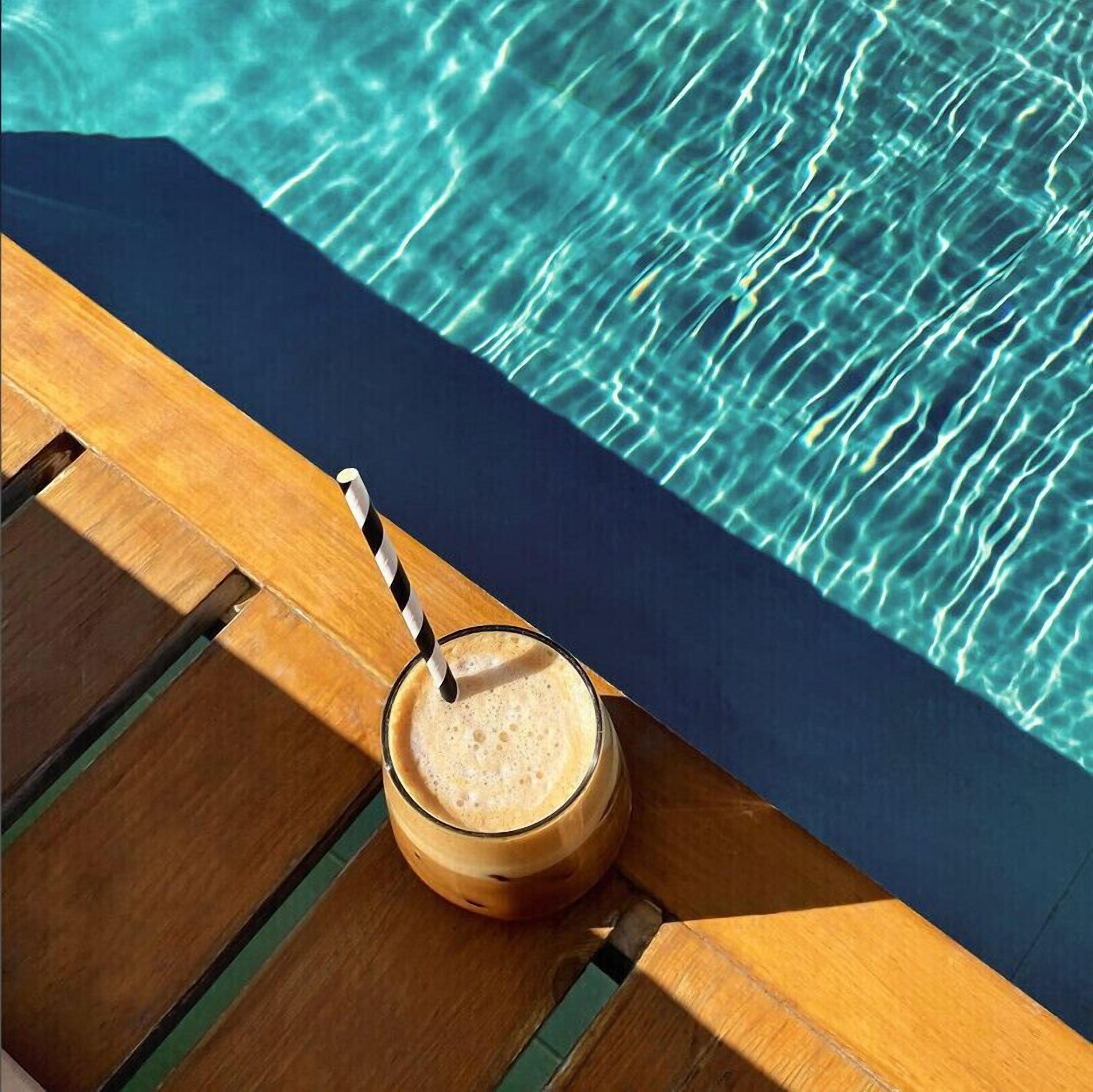 Un fauteuil de relaxation en bois, avec un verre contenant une boisson et une paille rayée, placé au bord d’une piscine à l’eau claire.