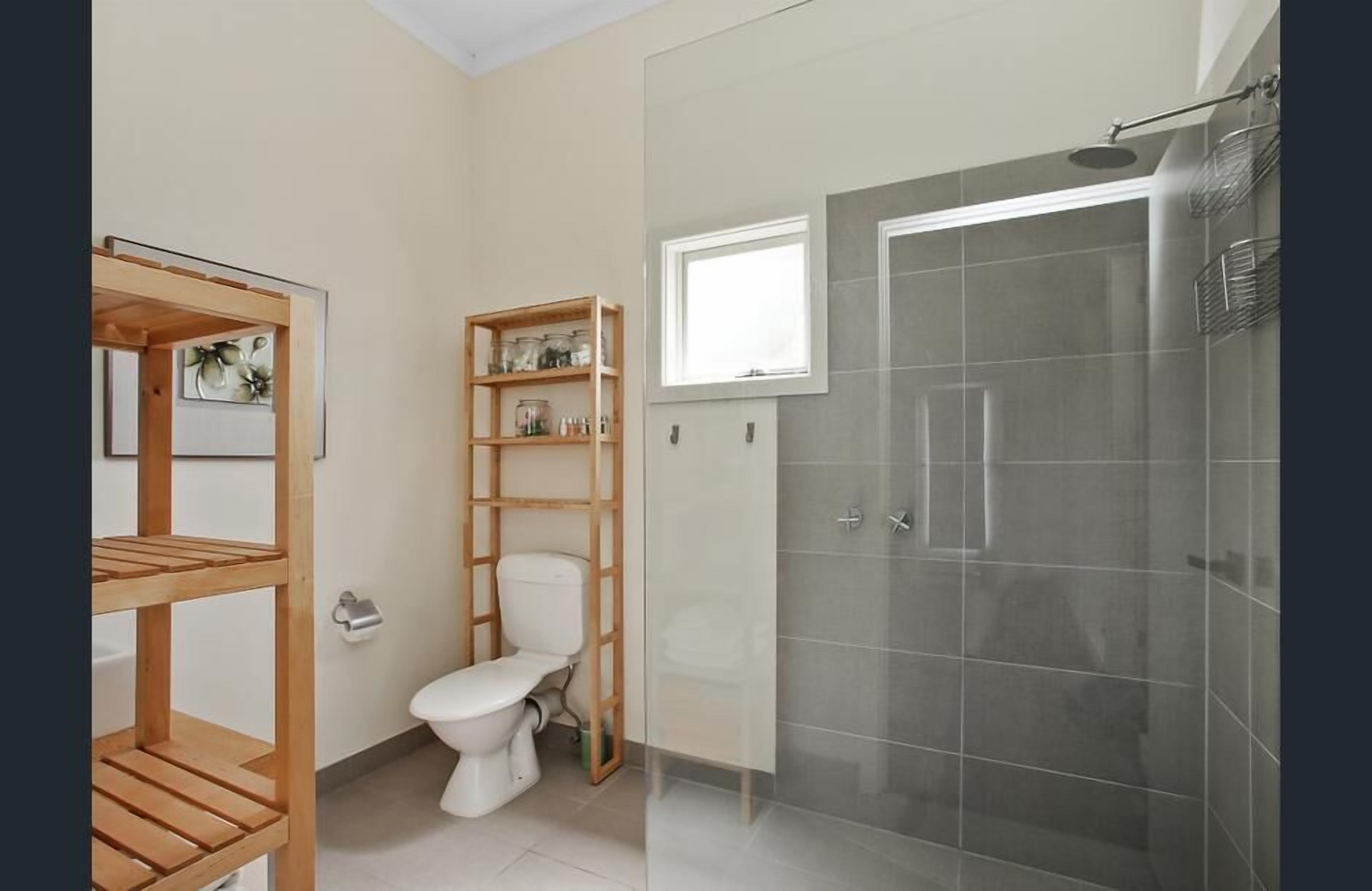 A bathroom with a toilet, wooden shelves, a shower with a glass door, and a window.