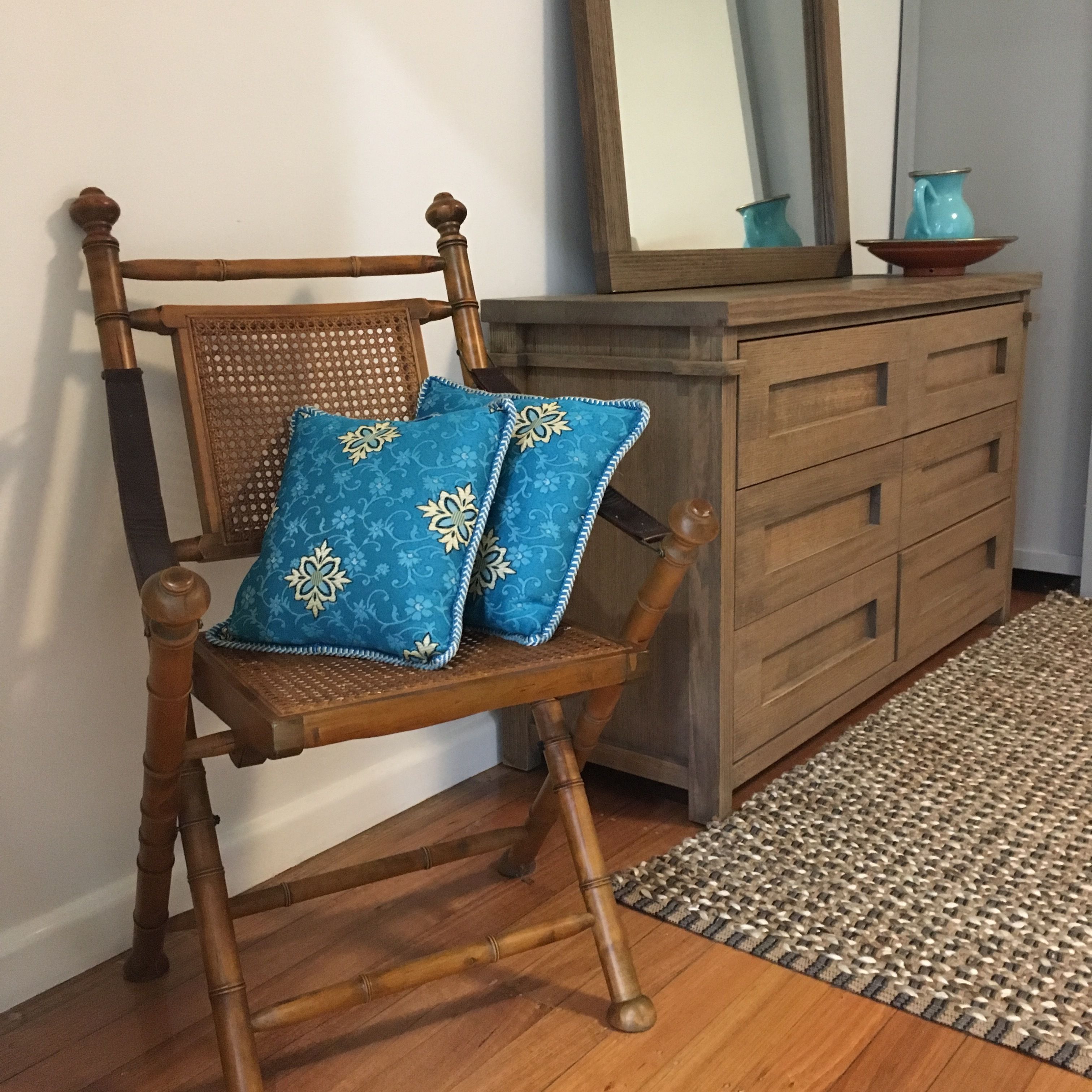 A wooden chair with blue patterned cushions, a wooden dresser with drawers, and a woven rug on a wooden floor.