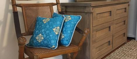 A wooden chair with blue patterned cushions, a wooden dresser with drawers, and a woven rug on a wooden floor.