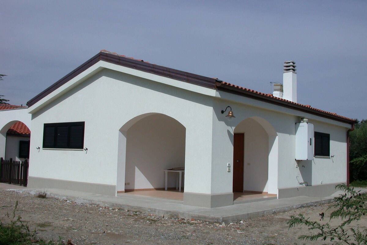 A single-story house with a tiled roof, a small porch with a table, and a bell on the wall.