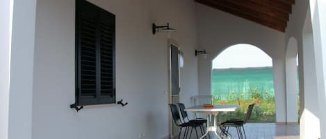 A covered patio with a table and chairs, a view of the sea, and a green garden in the background.