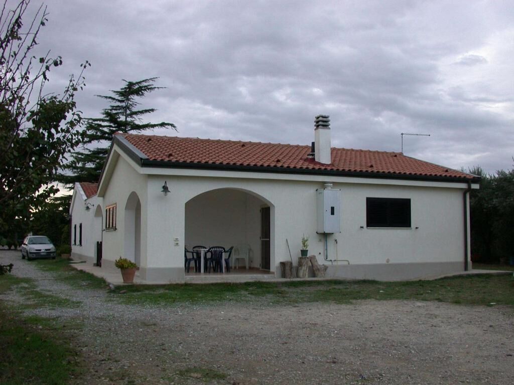 Un bâtiment d’un seul étage, doté d’un toit en tuiles, d’un patio couvert avec des sièges extérieurs et d’un garage visible.