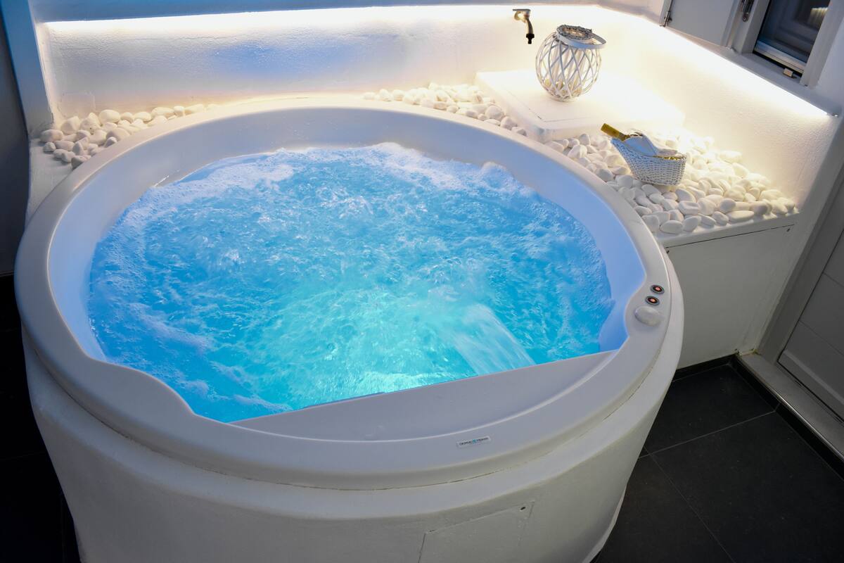 A hot tub with a white interior, surrounded by white pebbles, and a small table with a decorative item and a bottle.
