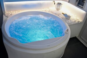 A hot tub with a white interior, surrounded by white pebbles, and a small table with a decorative item and a bottle.