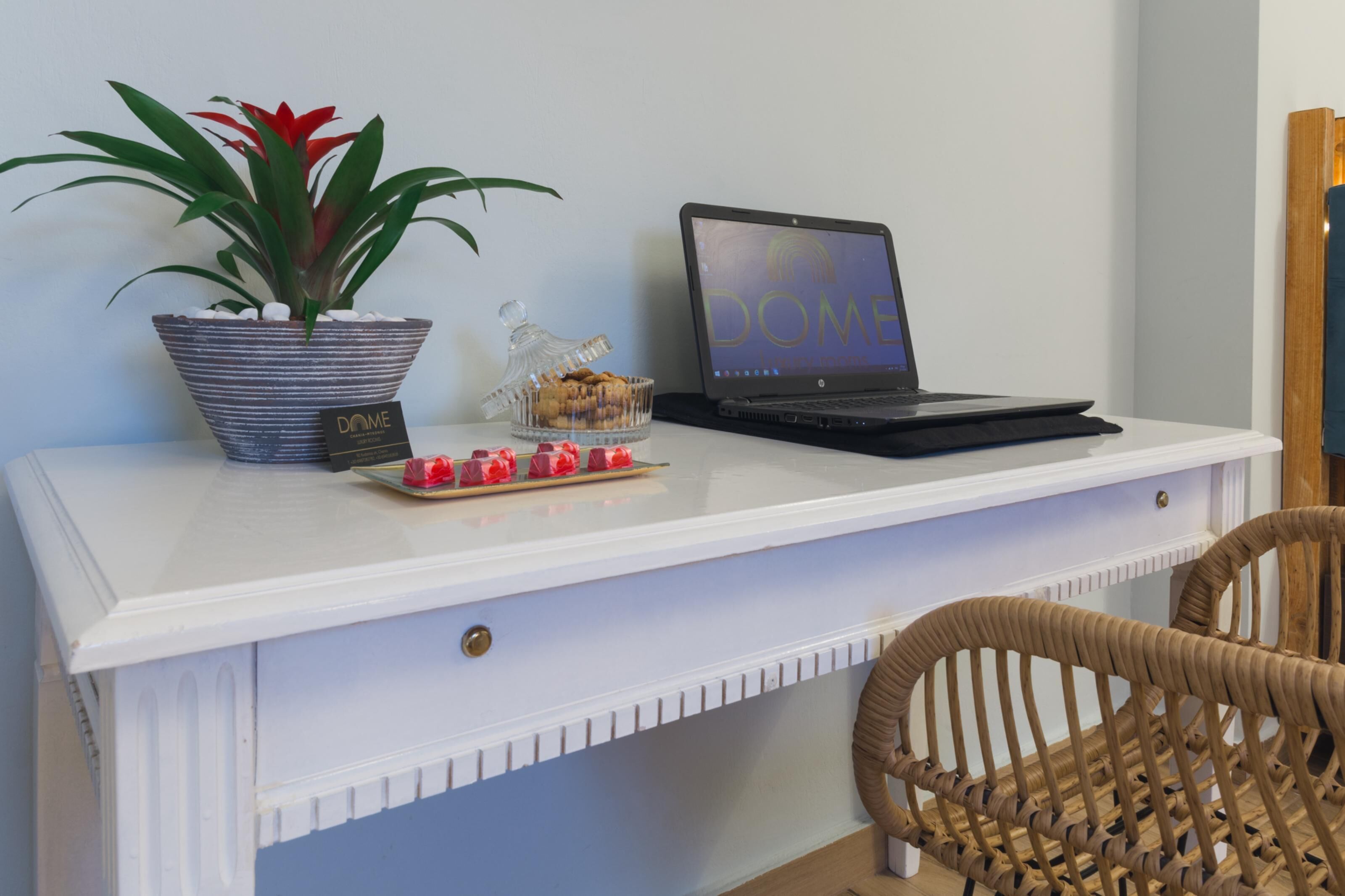 A white desk with a laptop, a potted plant, a glass container with cookies, and a wicker chair.
