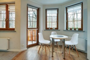 A room with a round table and four white chairs, large windows with wooden frames, and a radiator on the wall. - EverySky Lesny Dom (Karpacz)