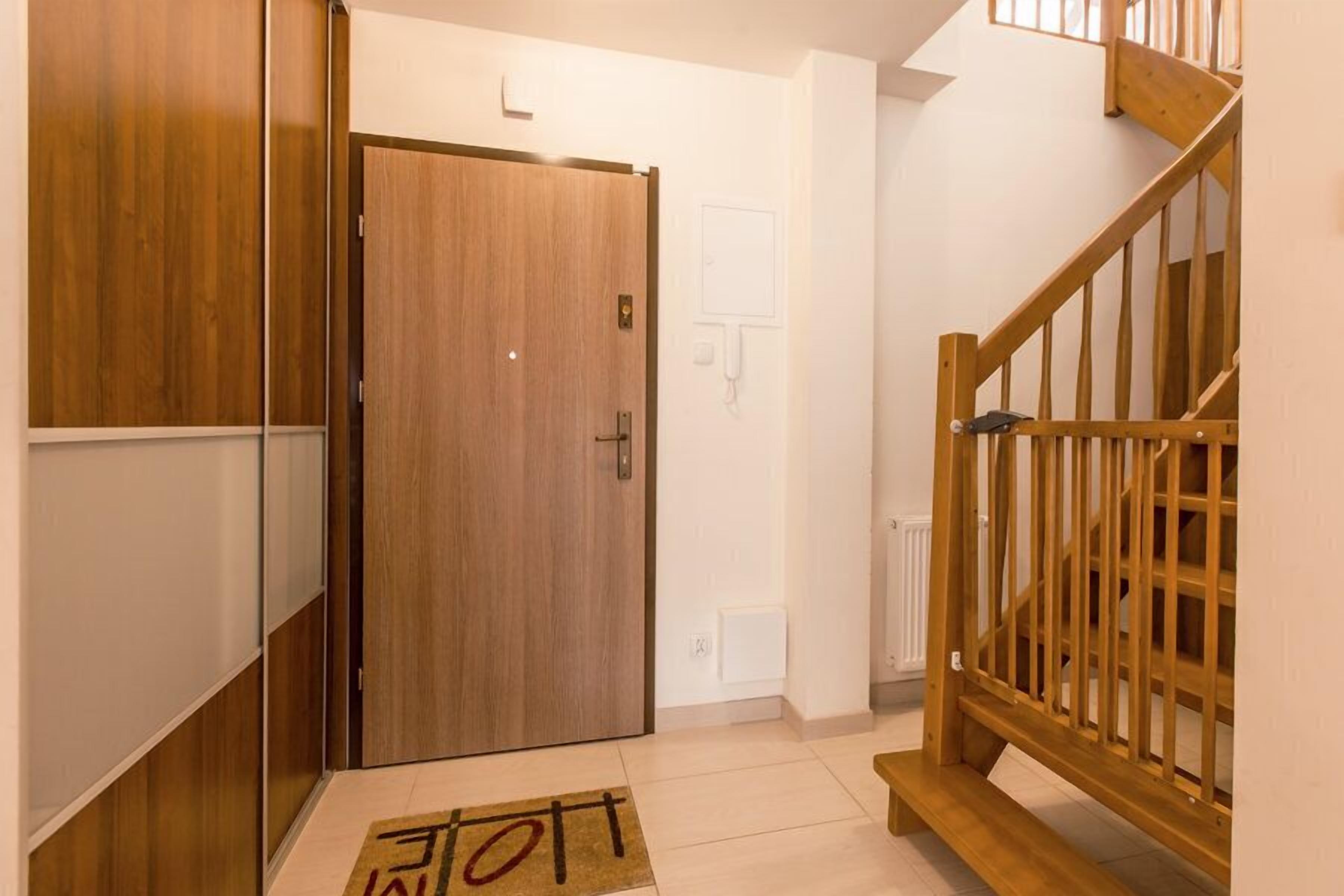 A wooden door with a handle, a white wall with a light switch, and a wooden staircase with a railing.