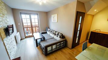 A cozy living area with a sofa, a small table, and a glass-top dining table. There is a window with a view of the outside, and a door leading to another room.