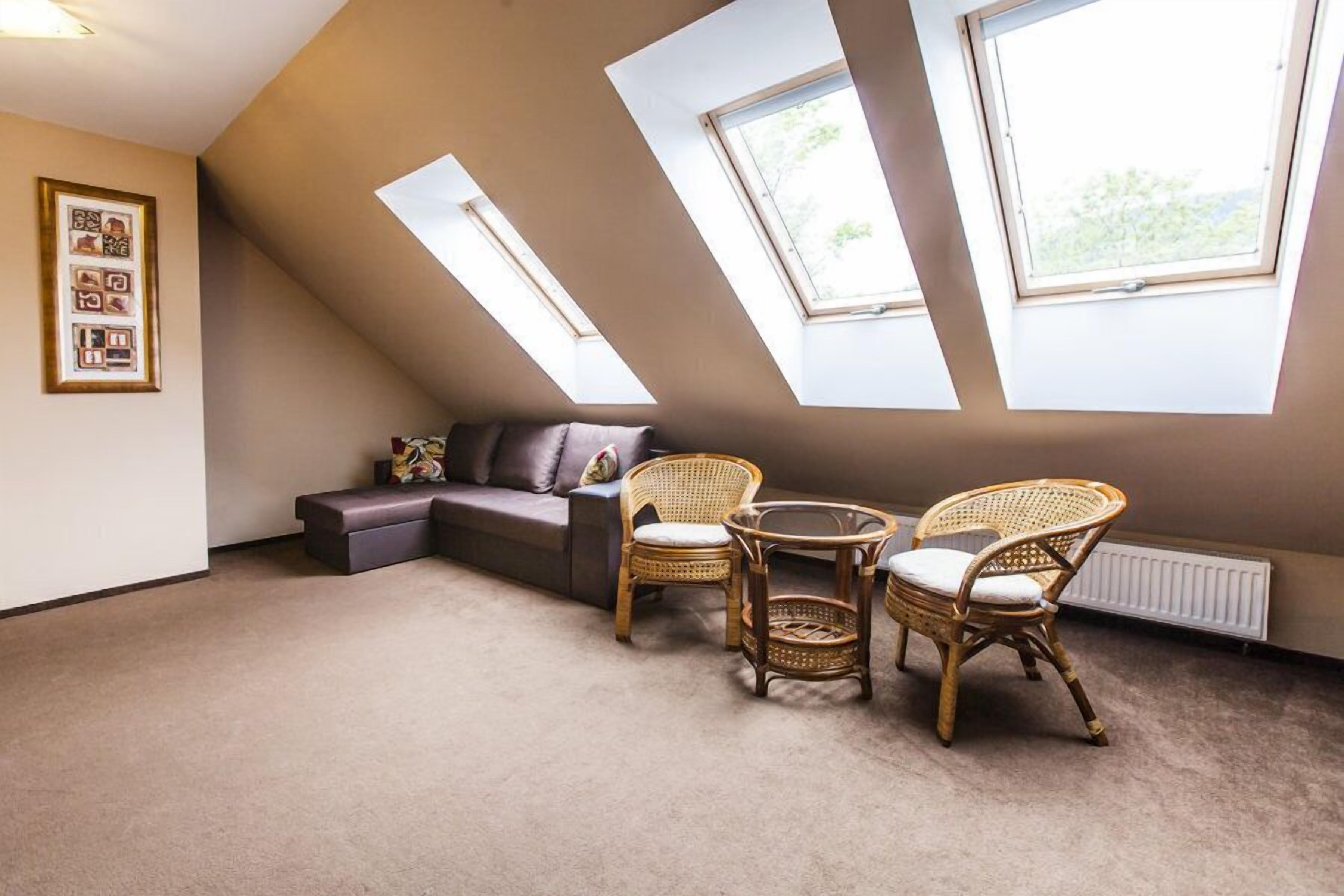 A room with a sloped ceiling, two large windows, a sofa, two wicker chairs, and a small table.