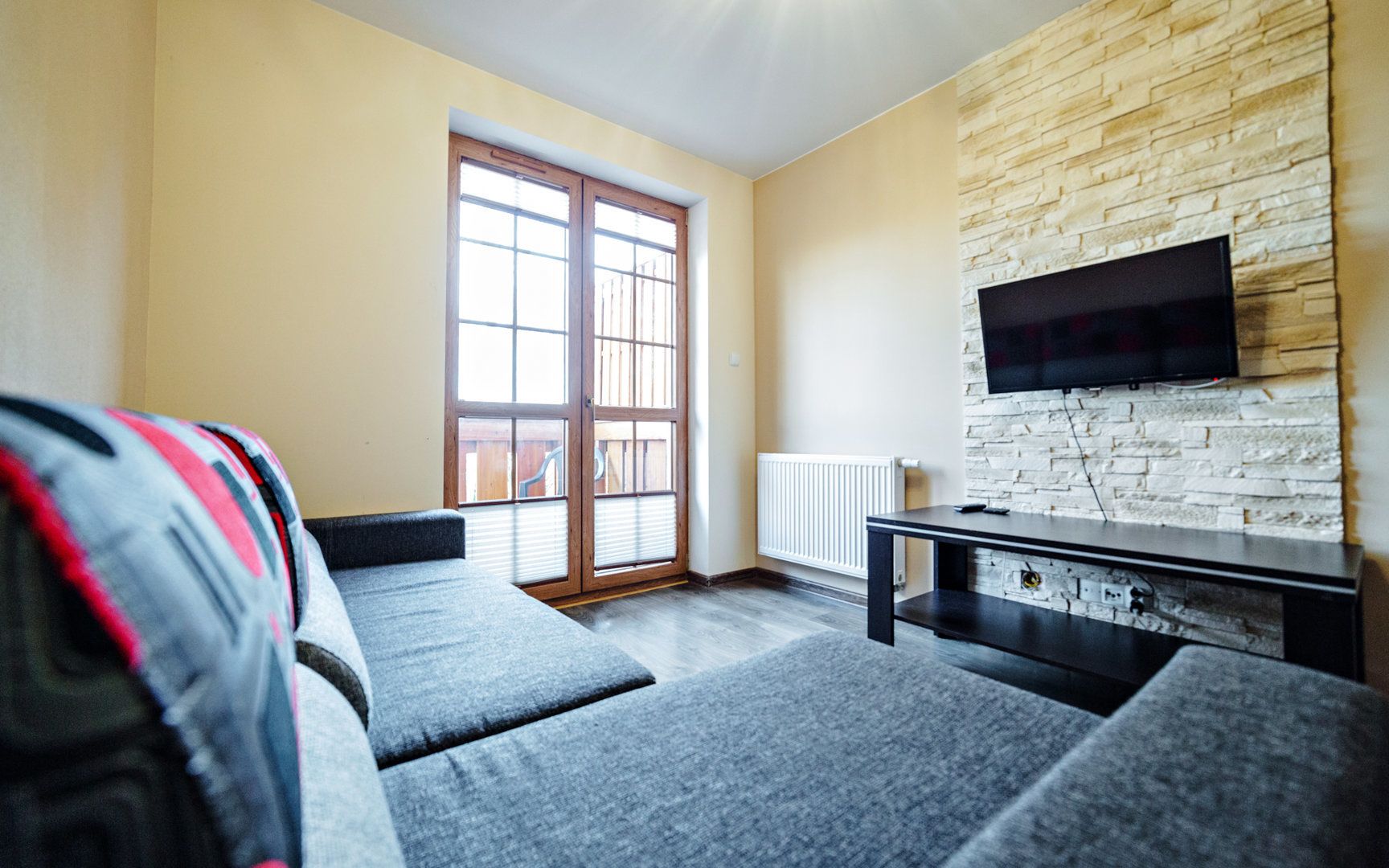 Een woonkamer met een bank, een televisie die aan een stenen muur is bevestigd, en een schuifdeur naar een buitenruimte.