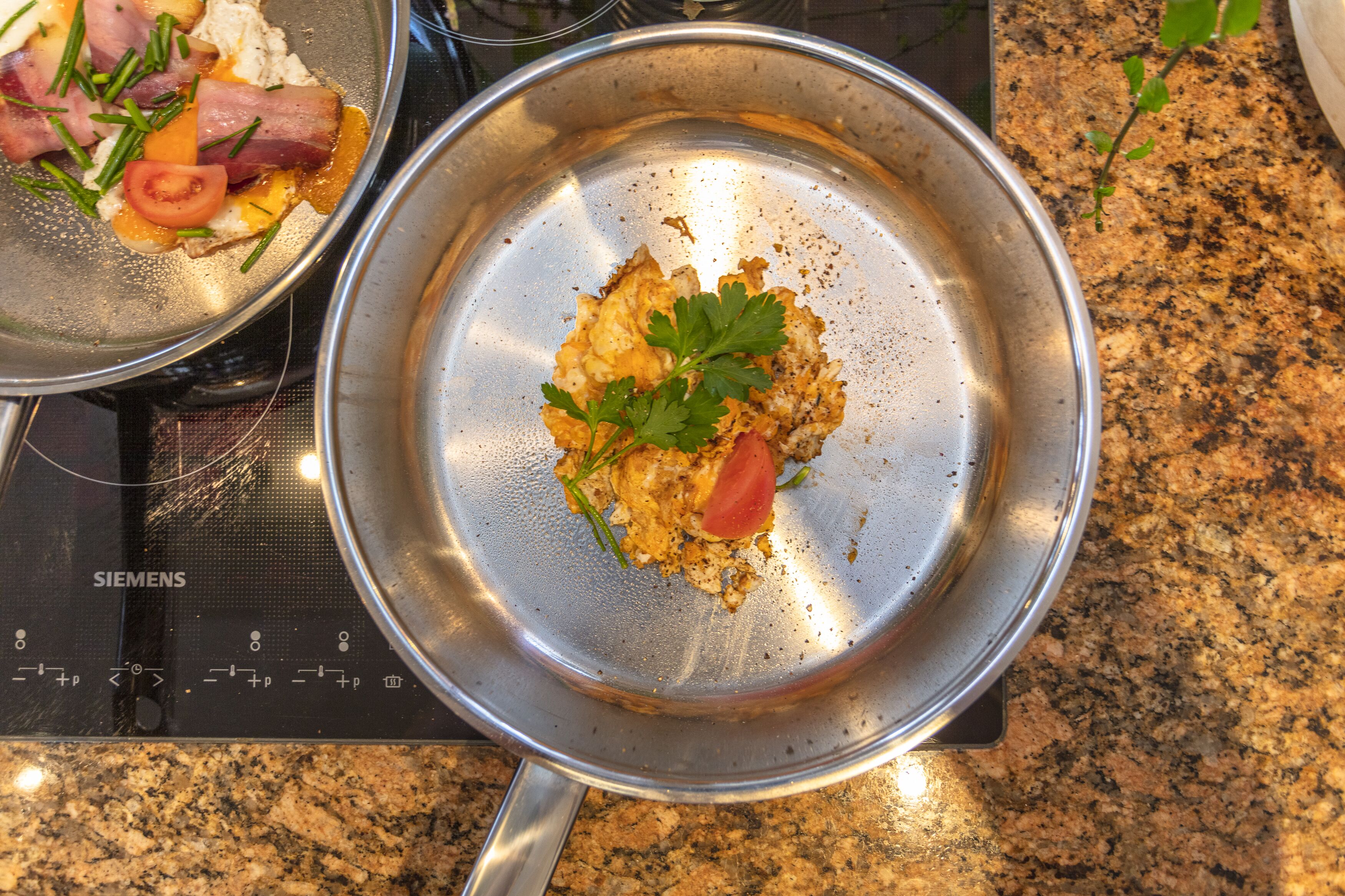 A stainless steel pan on a Siemens induction cooktop with scrambled eggs, a tomato, and green herbs.