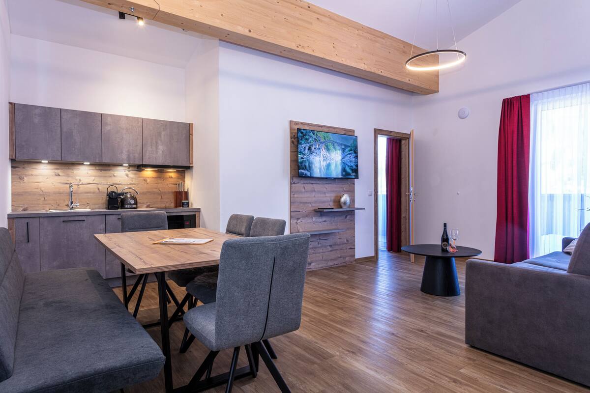 Een moderne woonruimte met een houten eettafel, gestoffeerde grijze stoelen, een bank en een wandgemonteerde flatscreen televisie.