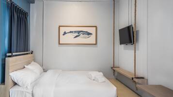 A neatly made bed with white linens, a framed whale artwork on the wall, a television mounted on a wooden shelf, and a rope hanging from the ceiling.