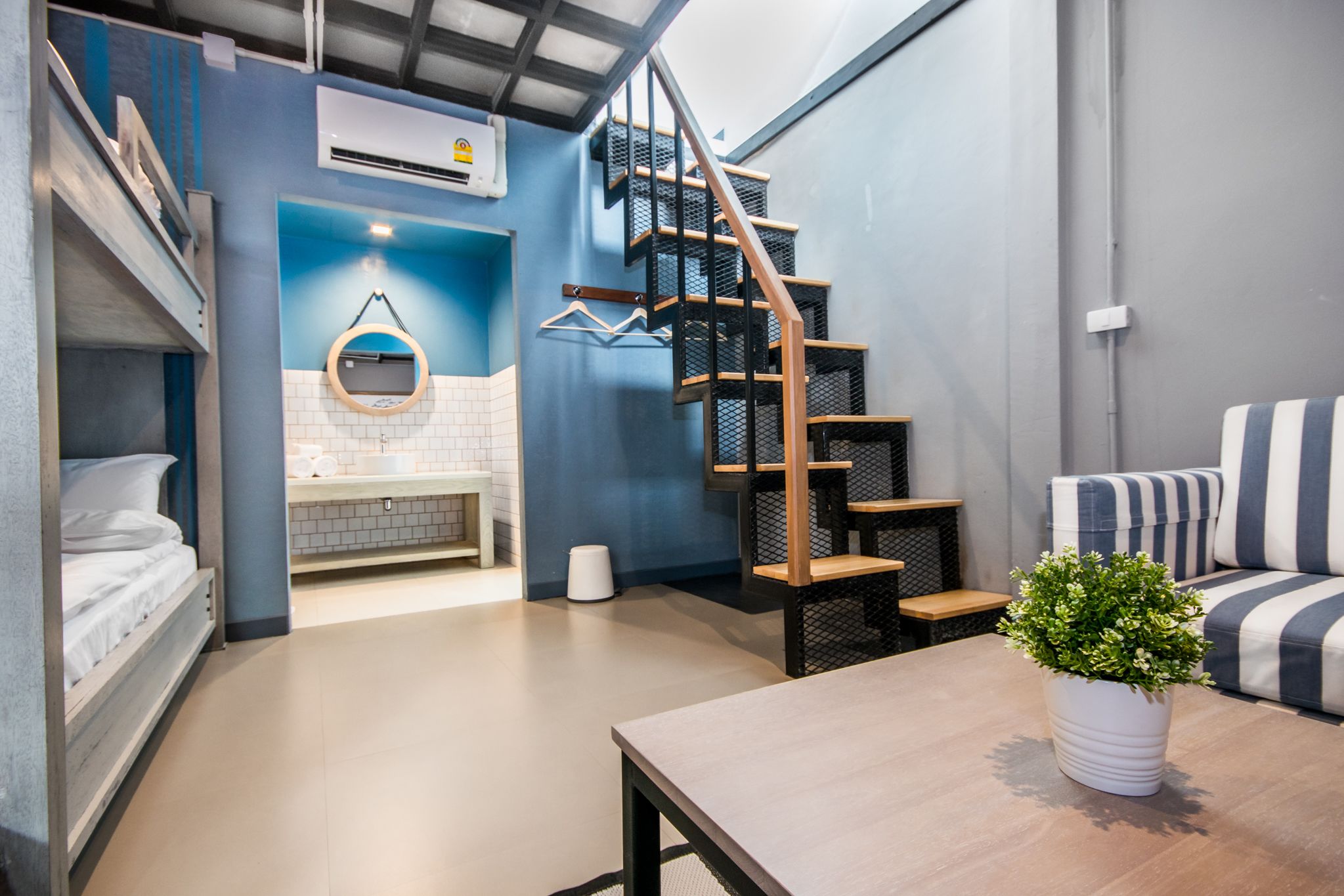 Un espacio habitable compacto con una cama litera, una pequeña cocina americana, una escalera y una zona de estar con un cojín a rayas.