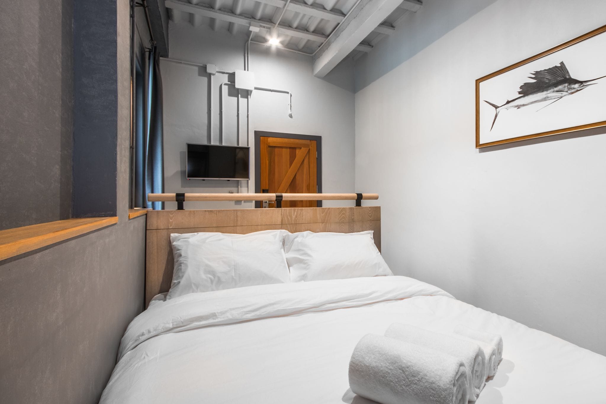 Moderni hotellihuone, jossa on puinen sängynpääty, valkoiset vuodevaatteet, seinälle asennettu televisio ja seinällä oleva kehystetty taideteos, jossa on kuvattuna kala.