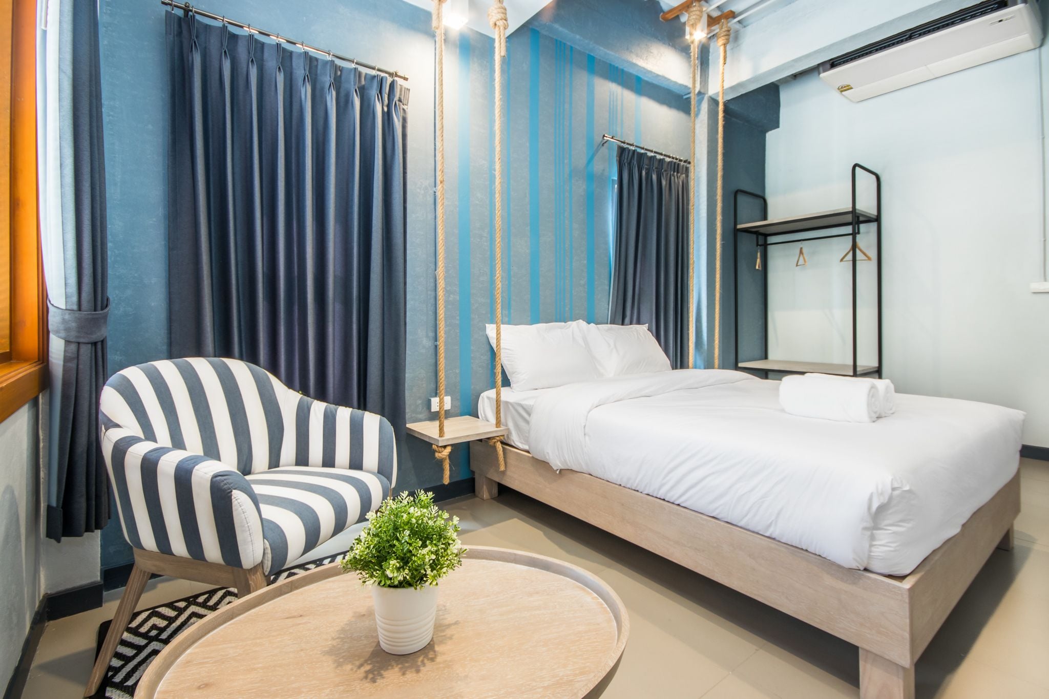 A modern hotel room with a bed, a striped armchair, a small table with a plant, and a hanging light fixture.