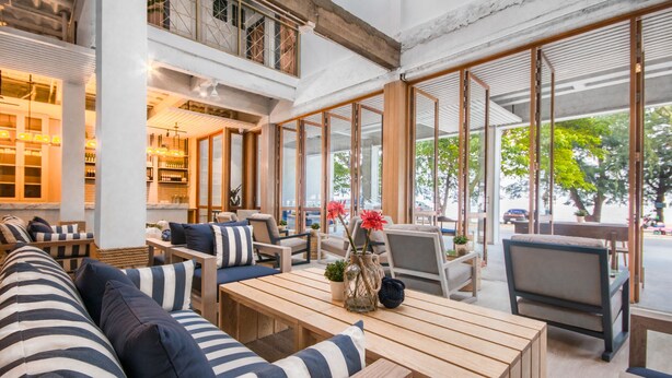 Un amplio salón interior con mesas de madera y sofás a rayas, grandes ventanales con vistas al exterior, y una zona de bar con estanterías llenas de botellas.