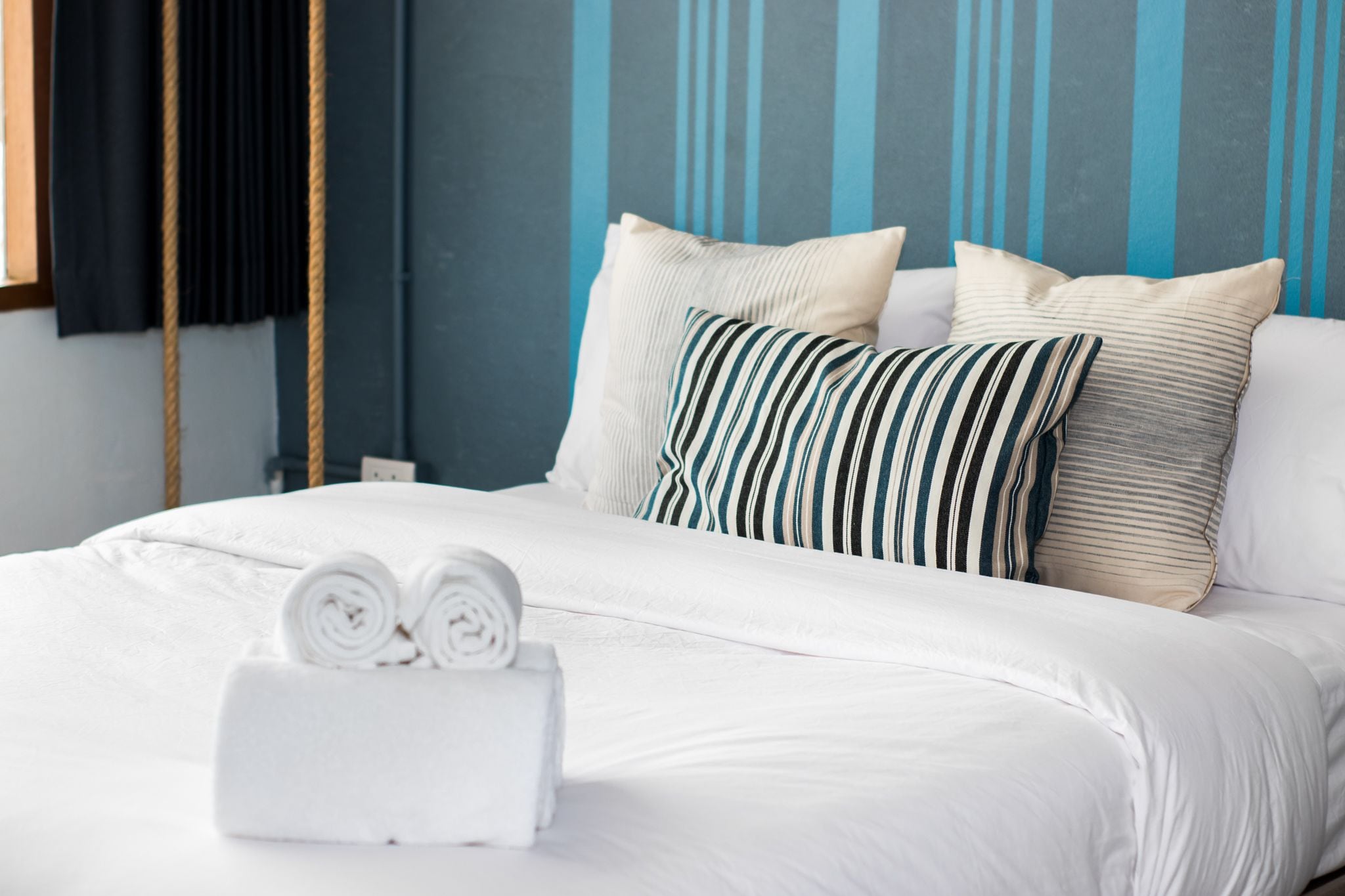 Una cama bien tendida con sábanas blancas y almohadas a rayas, una toalla blanca enrollada y una cortina oscura.