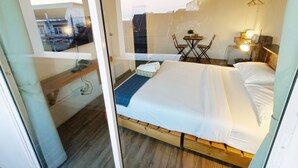 Un dormitorio con una cama, una pequeña mesa con sillas, un escritorio con lámpara y vistas a través de un ventanal arqueado.
