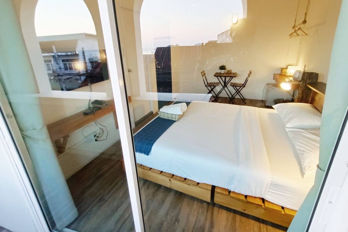 A bedroom with a bed, a small table with chairs, a desk with a lamp, and a view through an arched window.