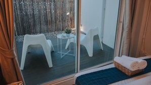 Habitación con cama, ventana y vistas a un espacio exterior con valla de bambú, dos sillas blancas y una pequeña mesa.