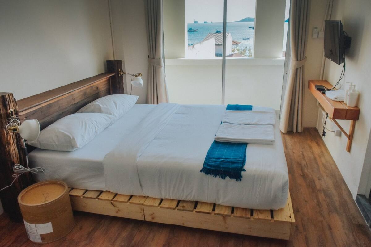A single bed with a wooden headboard, a bedside table with a lamp, a small wooden shelf with toiletries, and a view of the sea through a window.
